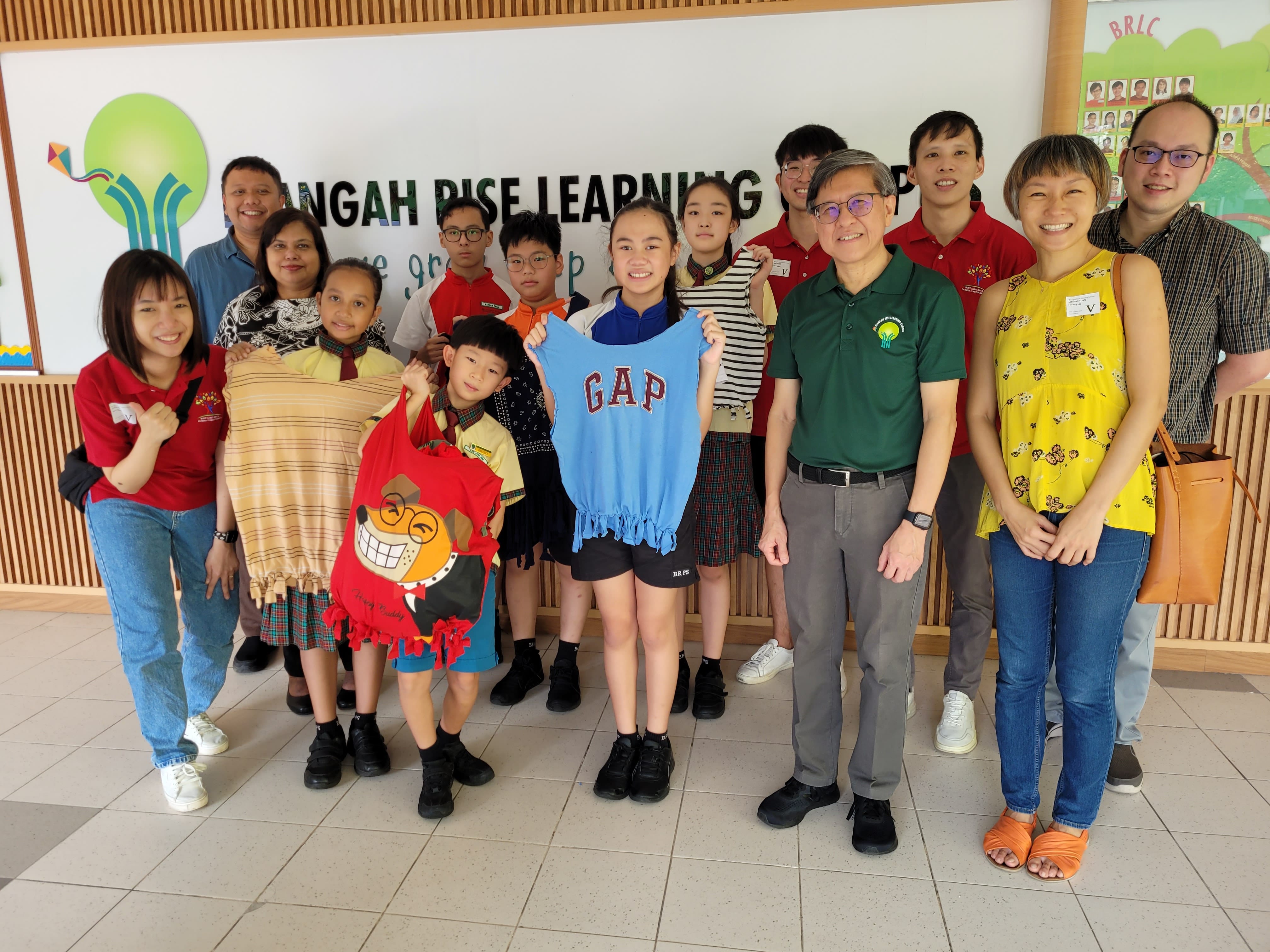 Group of adults and children posing indoors at Buangah Rise Learning Center, with kids holding handmade garments.